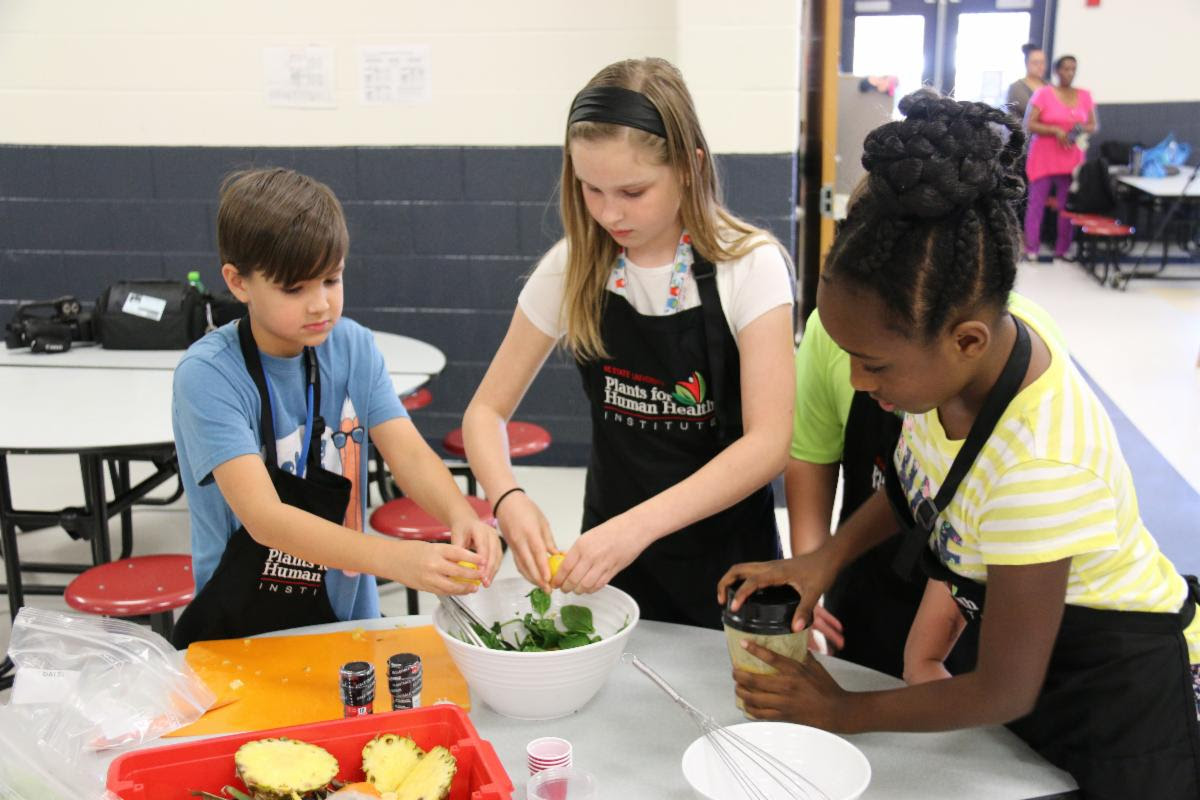 three elementary-aged students wearing aprons and preparing a salad together