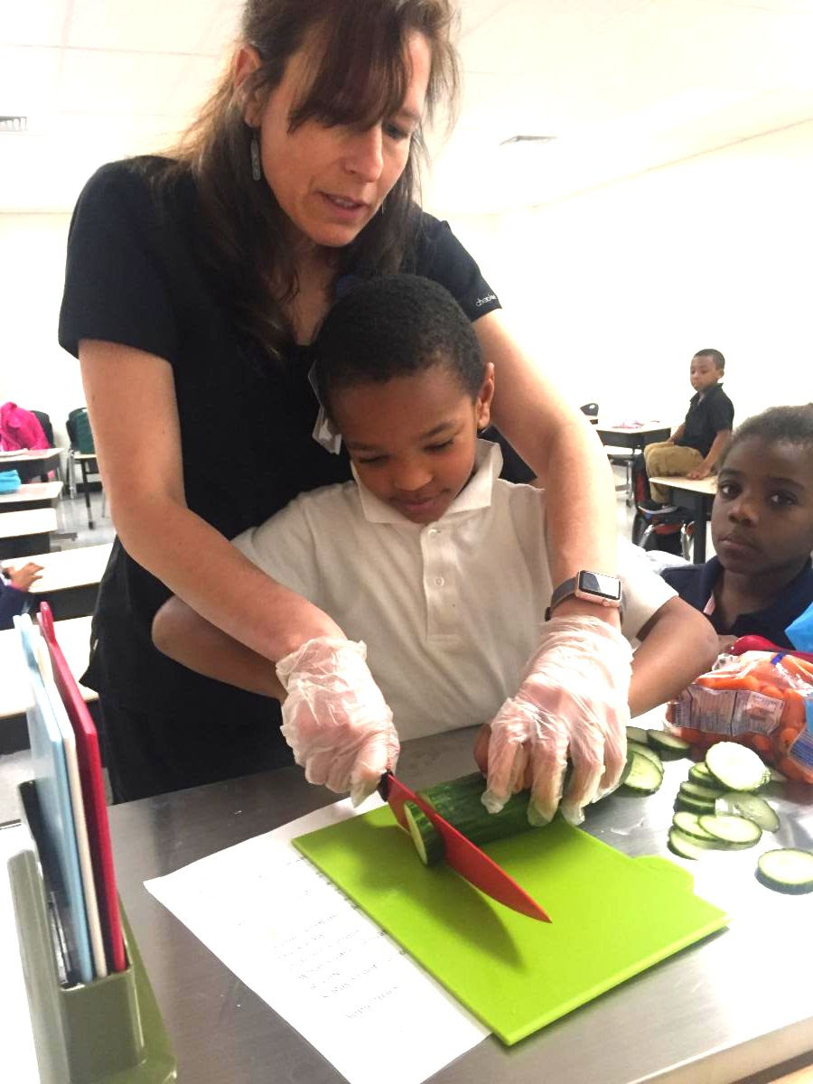 a teacher helping an elementary-aged student learn knife skills while slicing a cucumber on a cutting board