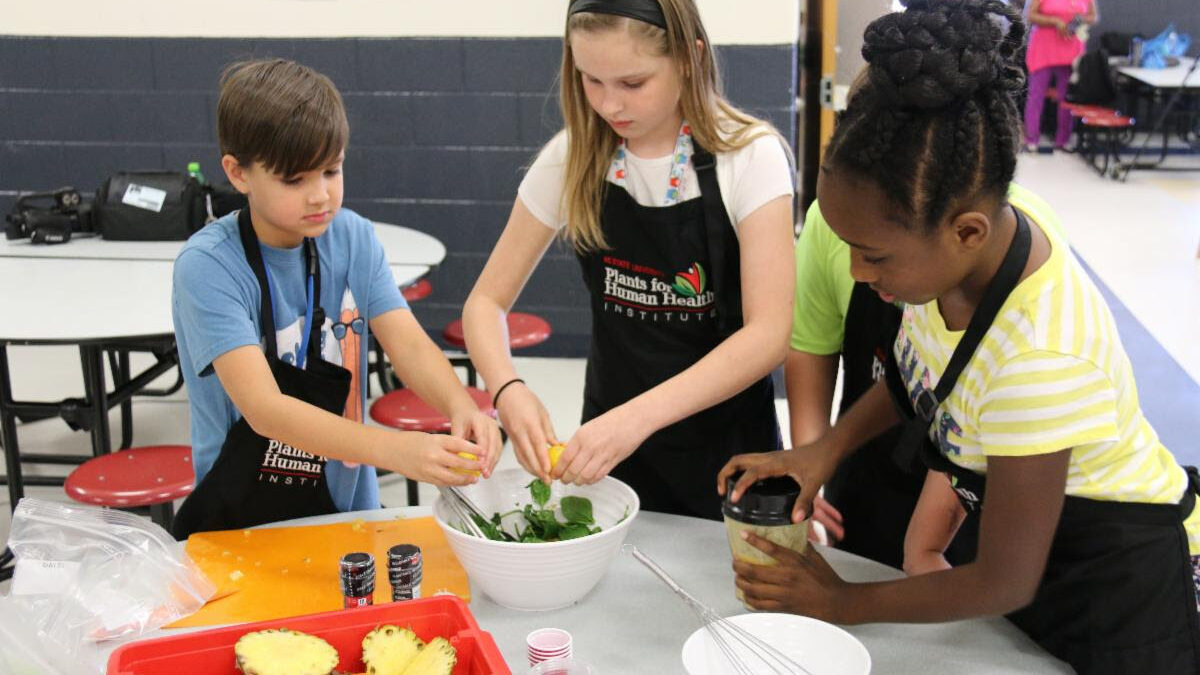 three elementary-aged students wearing aprons and preparing a salad together